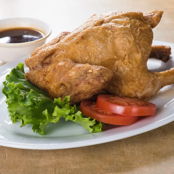 Whole roasted chicken on a white plate with lettuce, tomato slices, and a side of sauce.