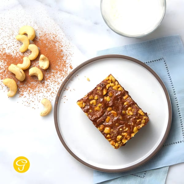 Goldilocks' cashew brownie on a white plate, with cashews, cocoa powder, and milk.